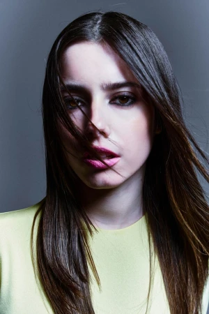 Close-up portrait of a young woman with long dark hair and strands across her face, wearing a yellow top against a grey studio background