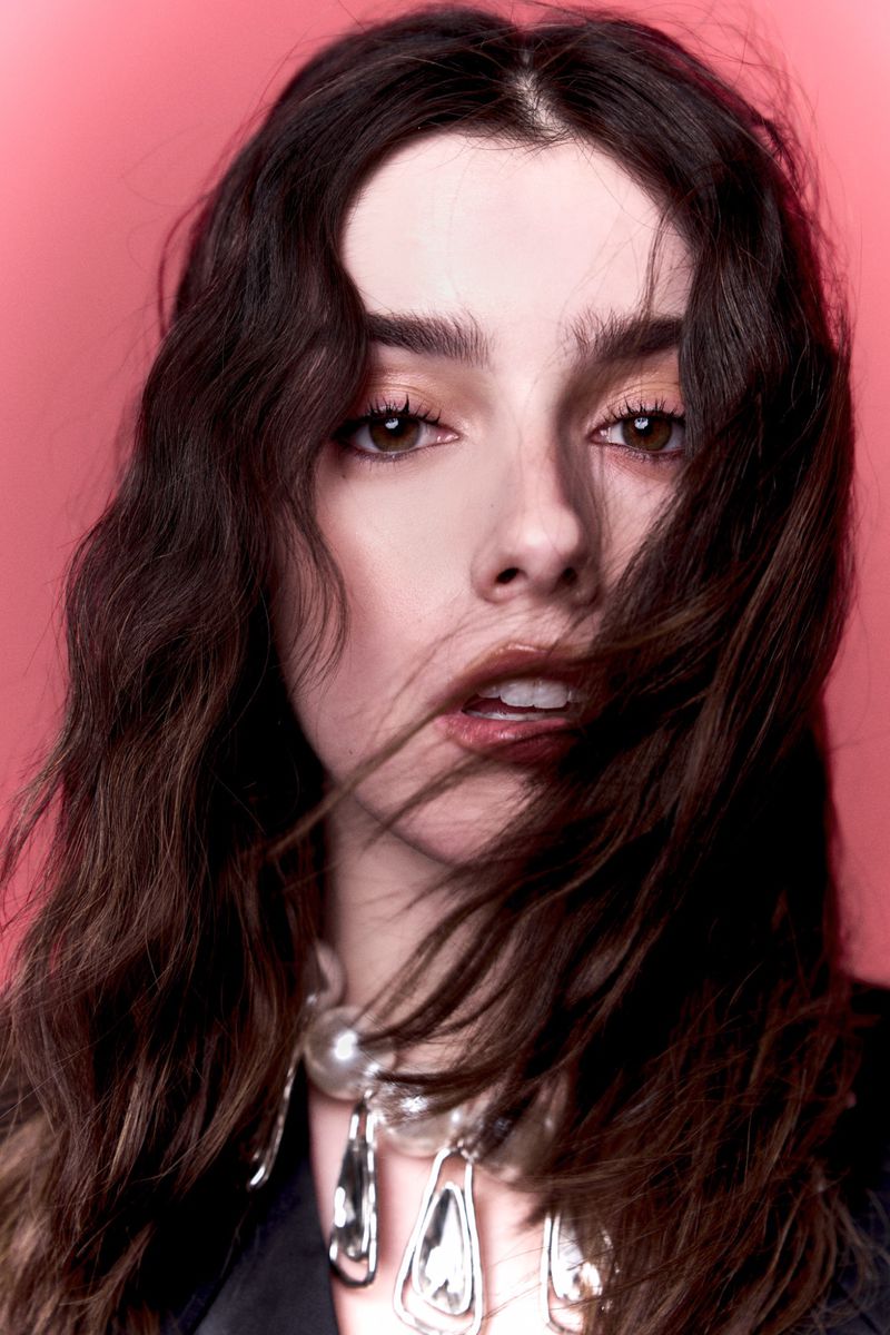 Portrait of a stylish adult woman with brunette wavy hair wearing a statement necklace, posing against a pink studio backdrop.