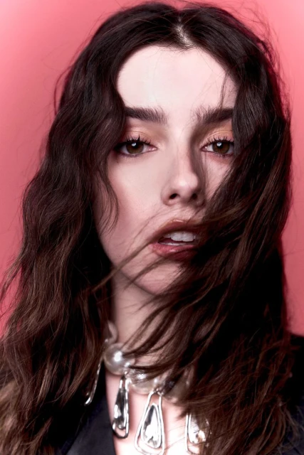 Portrait of a stylish adult woman with brunette wavy hair wearing a statement necklace, posing against a pink studio backdrop.