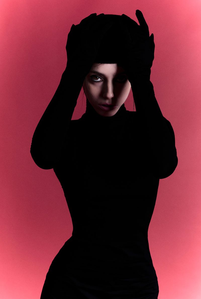 Madalena Aragão posing in a black high-neck silhouette with hands raised to her head against a glowing red studio backdrop.