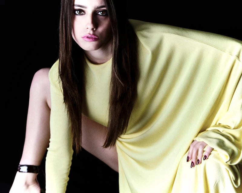 A young adult woman with long brown hair poses in a pale yellow long-sleeve shirt against a dark background in a studio shot.