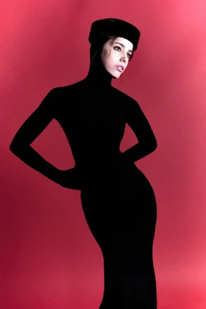 Madalena Aragão in a black high-neck dress and pillbox hat posing as a silhouette against a vibrant red backdrop.