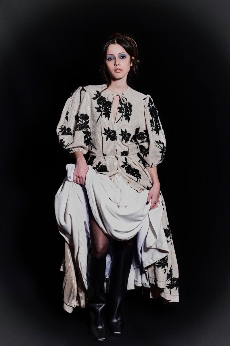 A woman poses in a beige floral dress, thigh-high boots, and holds her layered dress, against a stark black background in a studio shot.