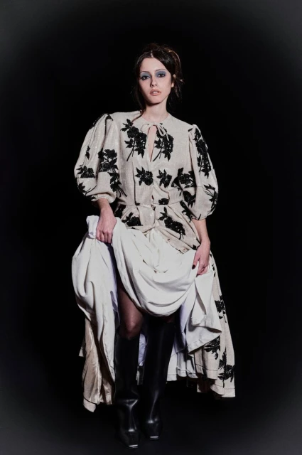 A woman poses in a beige floral dress, thigh-high boots, and holds her layered dress, against a stark black background in a studio shot.
