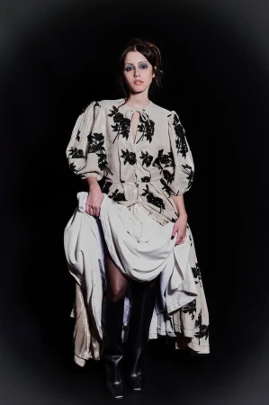 A woman poses in a beige floral dress, thigh-high boots, and holds her layered dress, against a stark black background in a studio shot.