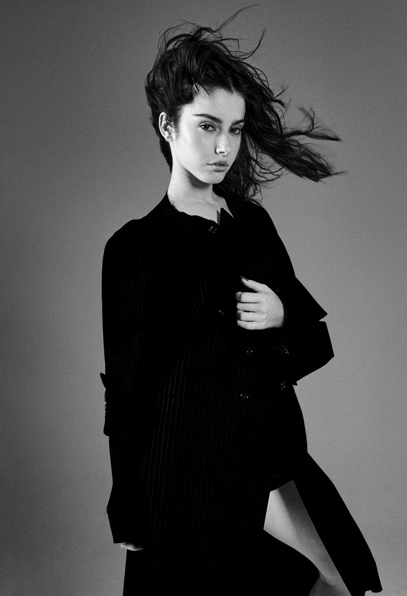 Black and white portrait of a young adult woman in a dark coat posing against a plain gray background in an indoor photo studio.