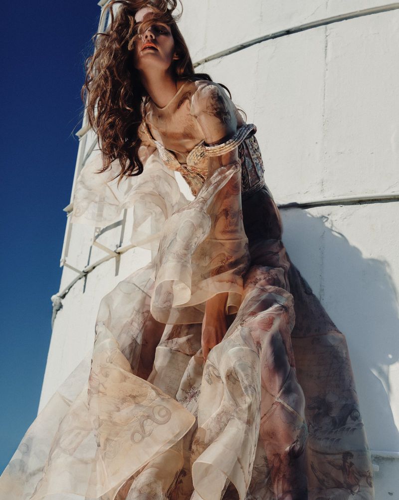 Adult female fashion model poses in beige gown with sheer overlay in outdoor daylight setting in front of a white building.