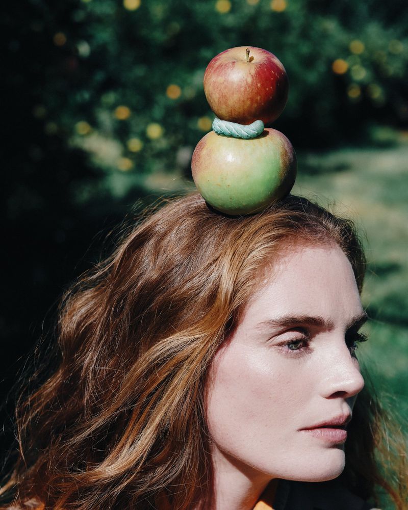 Woman in profile with auburn hair balancing two apples with green ring on her head in a green leafy outdoor setting.