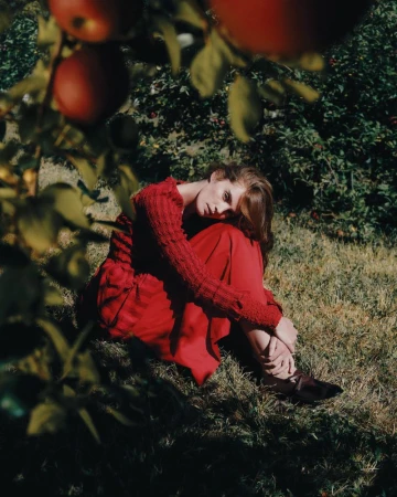 A young woman in a red dress sits in the grass beneath an apple tree, surrounded by green foliage and sunlight.