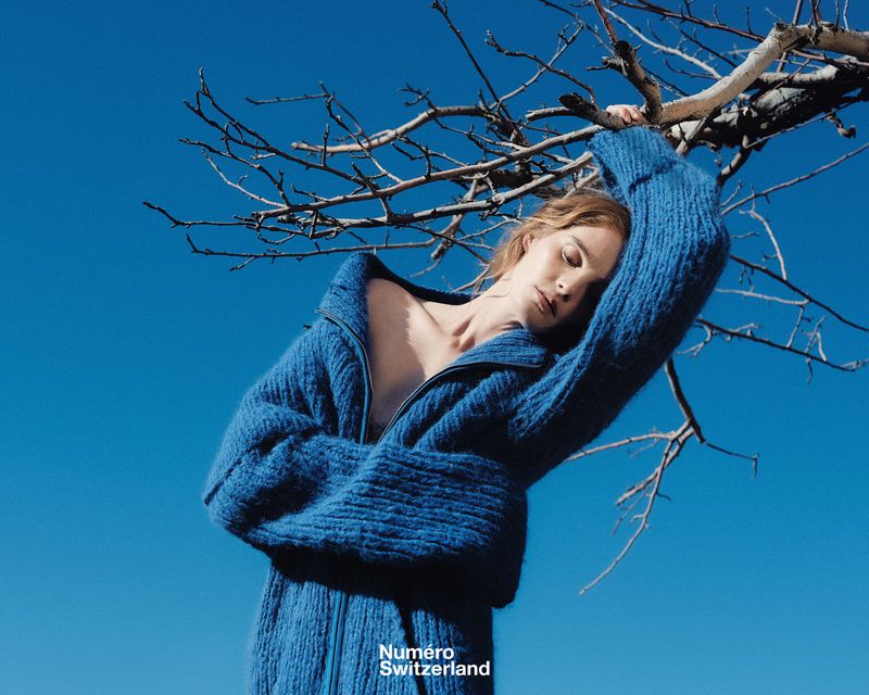 A woman with blond hair wears a blue sweater, leaning against a bare tree with a clear blue sky in the background. Fashion editorial shot.