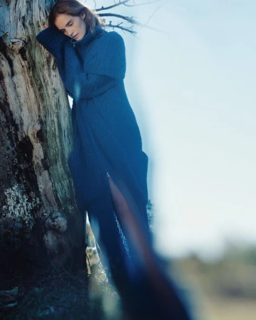 Adult woman in a long, knit blue dress leans against a tree in an outdoor setting with diffused lighting, creating a natural and serene scene.