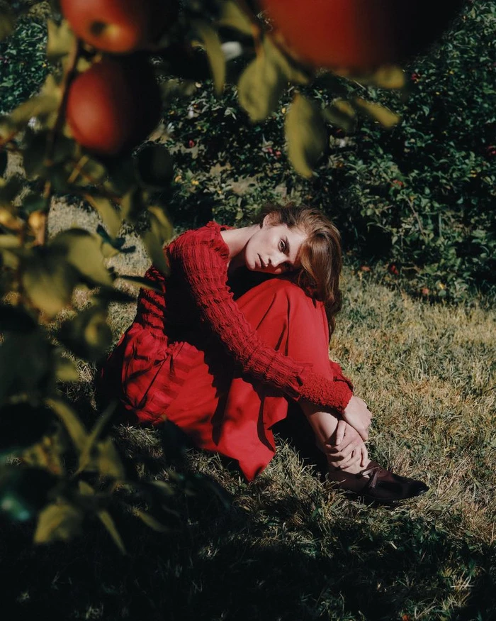 Model Alexina Graham wearing a red dress sitting in grass beneath an apple tree for Numero Switzerland by Lavina Solo