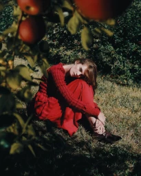 Model Alexina Graham wearing a red dress sitting in grass beneath an apple tree for Numero Switzerland by Lavina Solo