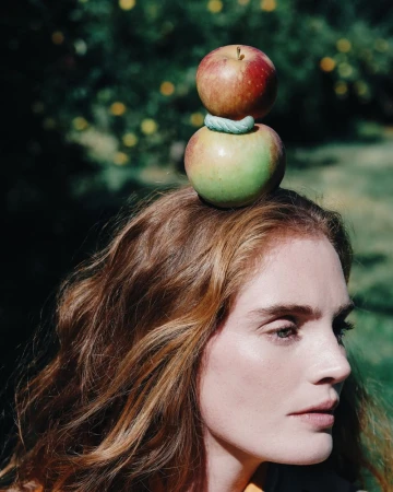 Alexina Graham with two stacked apples balanced on her head against a lush green outdoor background.