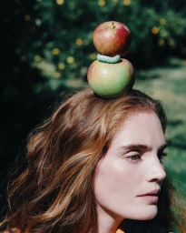 Alexina Graham with two stacked apples balanced on her head against a lush green outdoor background.