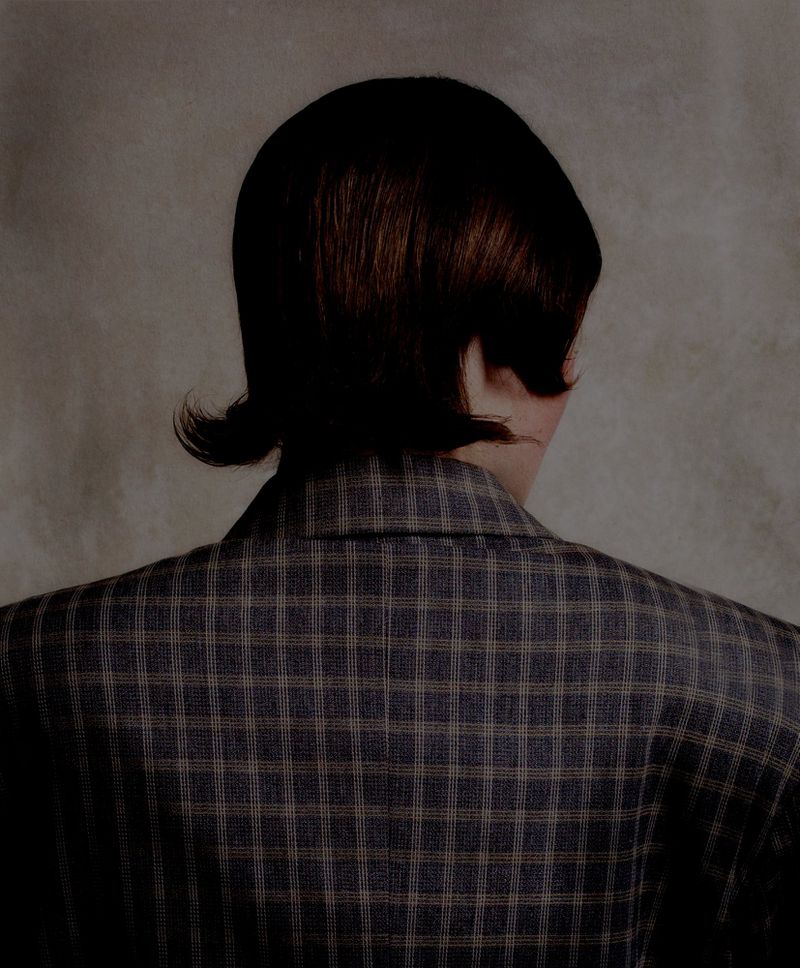 An adult with short brown hair from the back wearing a blue and brown plaid blazer jacket against a neutral background.