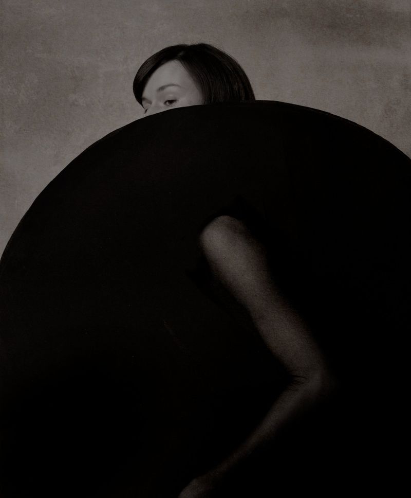 Monochrome portrait of a woman with a dark bob haircut, partially concealed by a large, rounded black object, against a textured background.