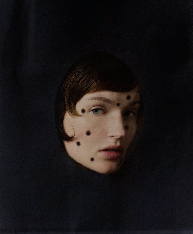 Artistic portrait of a woman with short brown hair and blue eyes against a dark background, face adorned with geometric black dots.