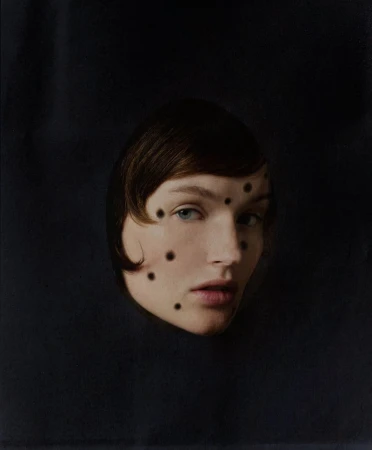 Artistic portrait of a woman with short brown hair and blue eyes against a dark background, face adorned with geometric black dots.
