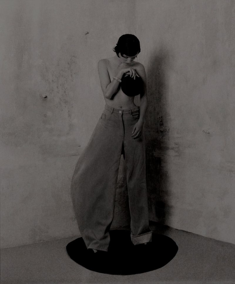 A person in wide-leg pants and a hat stands on a black platform, posing against a textured grey wall in a minimalist fashion image.