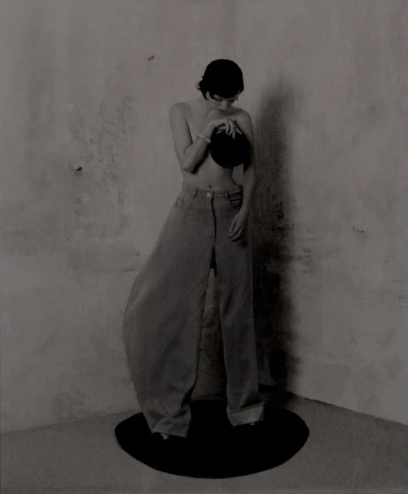 A person in wide-leg pants and a hat stands on a black platform, posing against a textured grey wall in a minimalist fashion image.