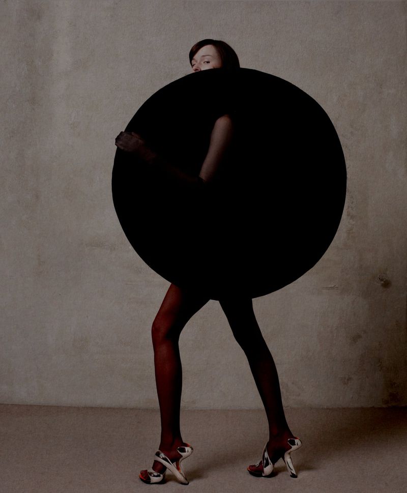 A slender woman in a black circle dress walks against a textured beige backdrop, accentuating unique high heels and dark red hosiery.