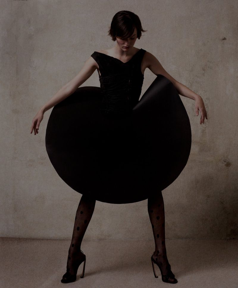 A female fashion model poses in a sculptural black dress with artistic styling and polka dot stockings against a neutral backdrop.