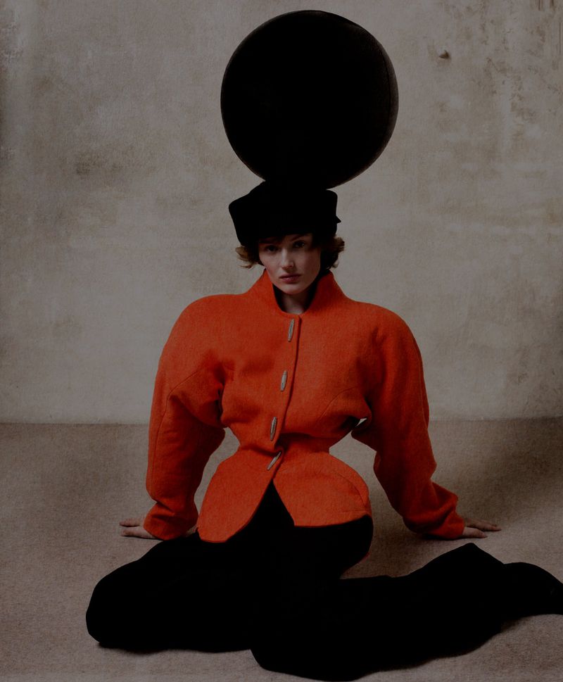 Fashion shot of a woman in an orange jacket and black pants, seated with an orb on her head, capturing an artistic, surreal aesthetic.