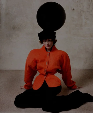 Fashion shot of a woman in an orange jacket and black pants, seated with an orb on her head, capturing an artistic, surreal aesthetic.