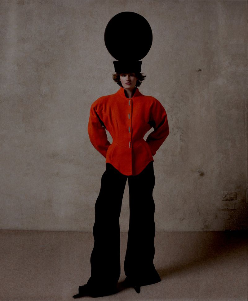 Full-length fashion shot of a female model wearing a red jacket, black pants, and a black hat with a large black disc on top against a neutral background.