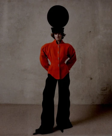 Full-length fashion shot of a female model wearing a red jacket, black pants, and a black hat with a large black disc on top against a neutral background.