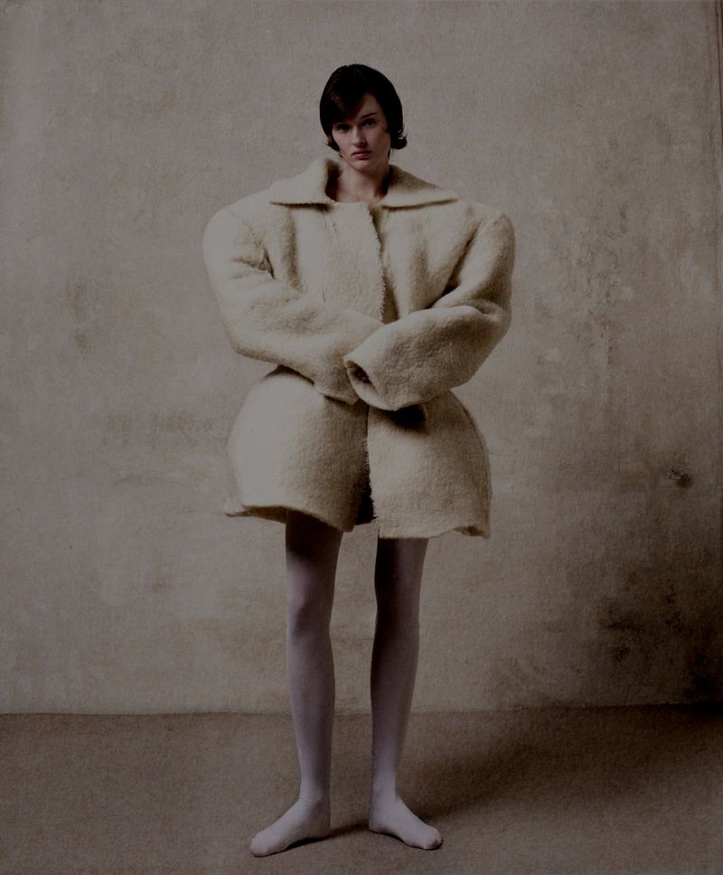 A woman poses in a cream-colored coat-like garment and white tights against a neutral backdrop in a studio fashion shot.