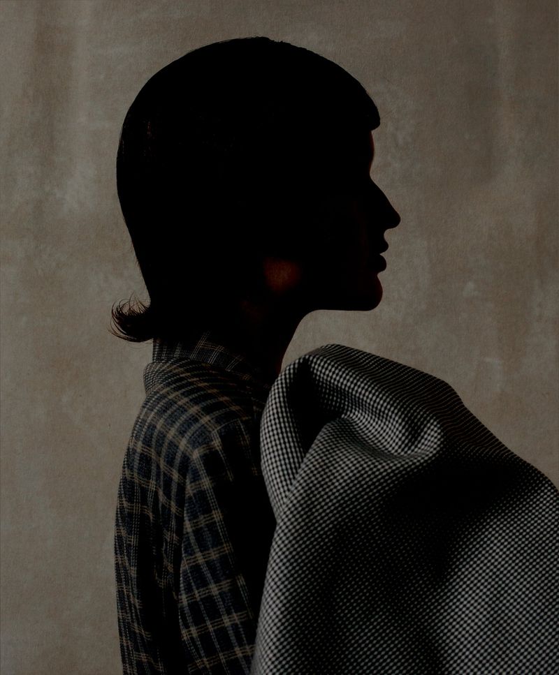 Profile of an adult female in a plaid shirt with a draped houndstooth jacket, set against a muted grey background in shadow.