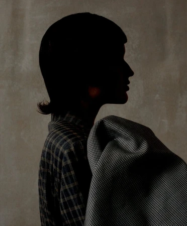 Profile of an adult female in a plaid shirt with a draped houndstooth jacket, set against a muted grey background in shadow.