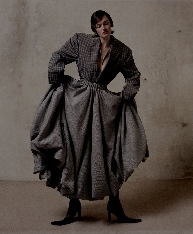 Woman in oversized plaid blazer and voluminous gray skirt stands indoors against a neutral backdrop in a fashion studio setting.