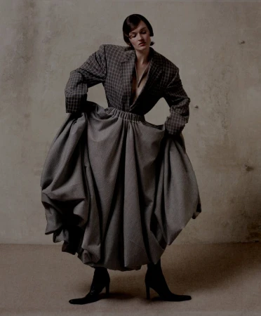 Woman in oversized plaid blazer and voluminous gray skirt stands indoors against a neutral backdrop in a fashion studio setting.
