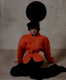 Model seated on the floor wearing a structured orange jacket and black trousers, positioned below a large black sphere.