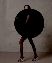 Model holding large black circular prop in front of torso against textured neutral concrete wall