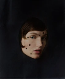 A close-up portrait of a woman with dark dots on her face framed by an oval opening in a dark black background.