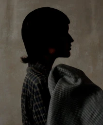 Profile silhouette of a model in a plaid shirt holding draped houndstooth fabric against a neutral textured wall.