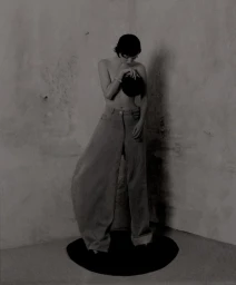 Model posing topless in wide-leg high-waisted trousers holding a black disc in a textured concrete studio corner.