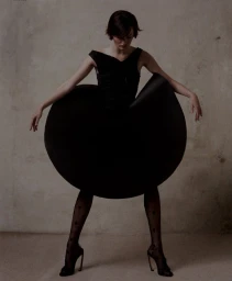 Model posing in a sculptural black circular disc dress, patterned tights, and stiletto heels against a neutral backdrop.