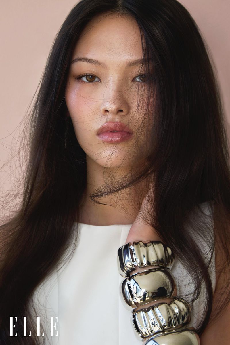 Close-up portrait of an Asian woman with long dark hair wearing white sleeveless top and large silver bangle, posing in a photo studio.