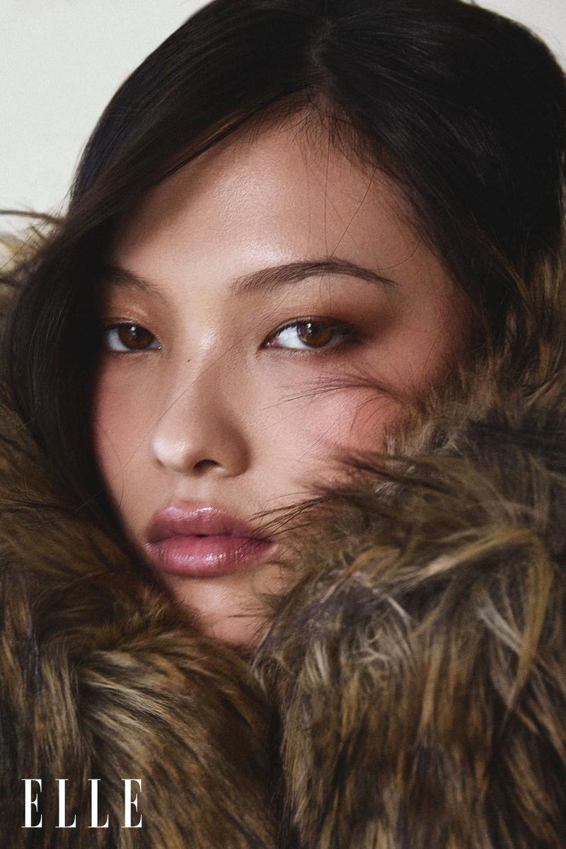 Close-up portrait of a young Asian woman with brown eyes and dark hair, partially framed by a brown faux fur collar and the ELLE logo.