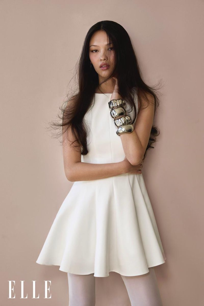 Model May Li Wescott wearing a white sleeveless flared mini dress and chunky silver cuff bracelets against a warm studio backdrop.