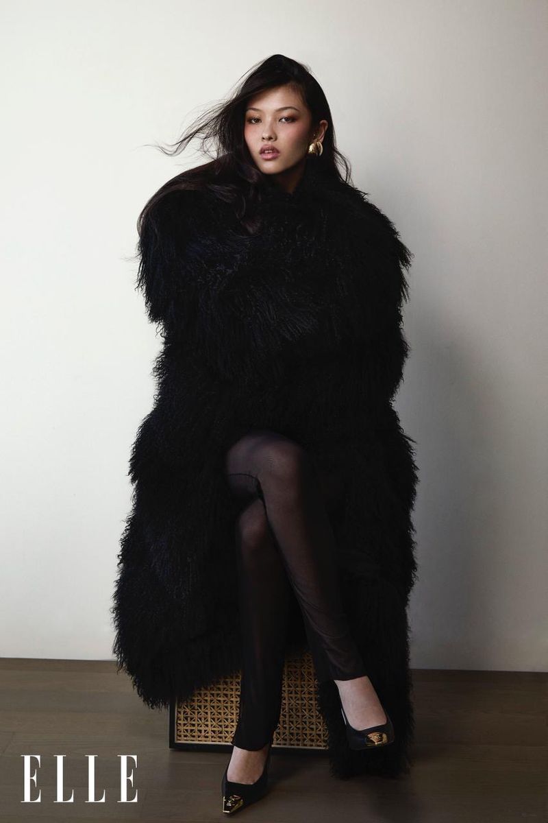 Portrait of a young woman in a long, shaggy black coat, leggings, and black shoes, sitting on a rattan stool against a white wall.