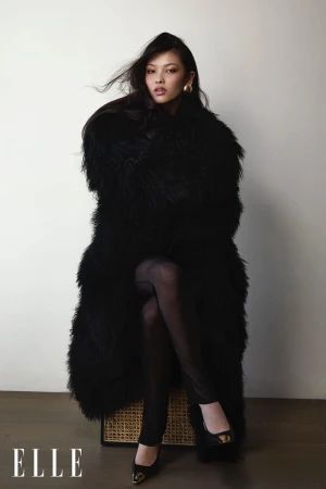 Portrait of a young woman in a long, shaggy black coat, leggings, and black shoes, sitting on a rattan stool against a white wall.