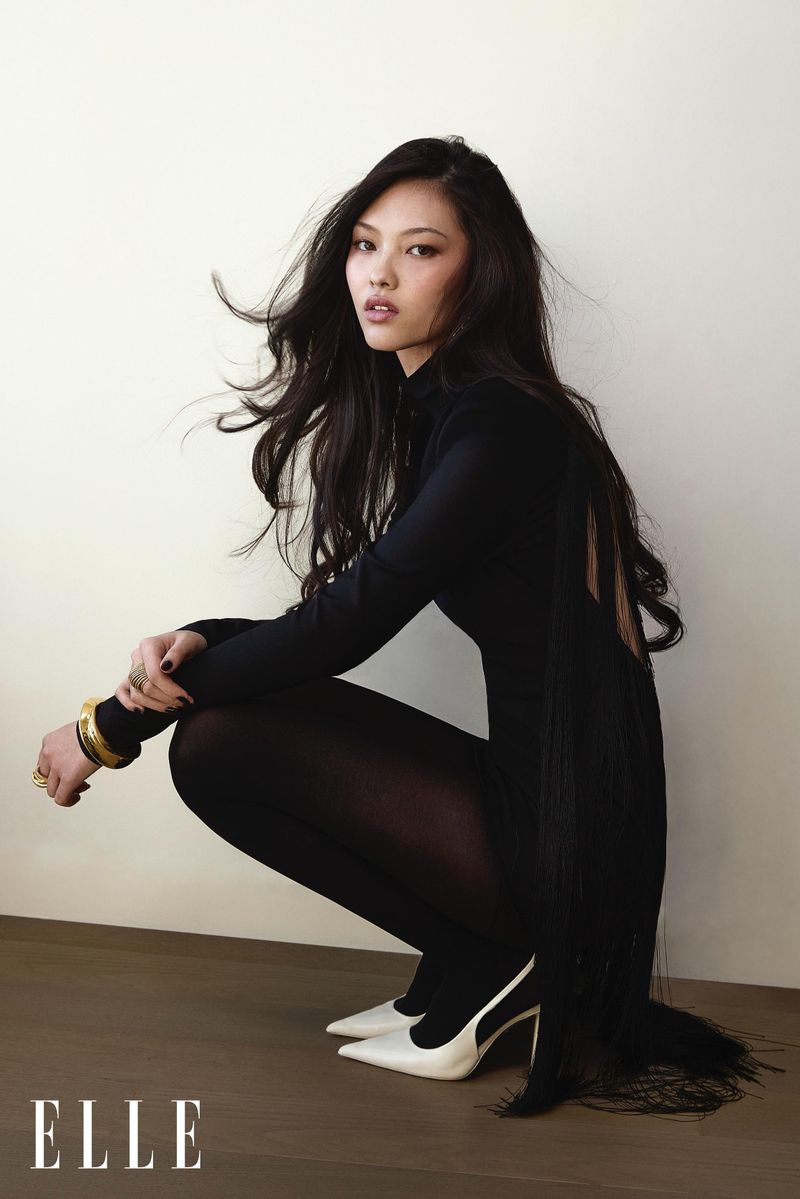 Portrait of a crouched Asian woman with long dark hair, wearing a black dress, matching tights and white heels, shot in a photo studio.