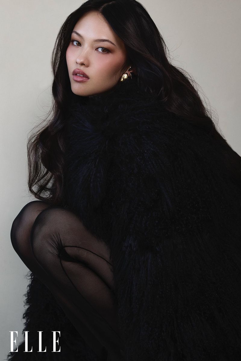 Close-up portrait of an Asian woman in dark clothing, kneeling in front of a neutral background. She has long hair and gold earrings.