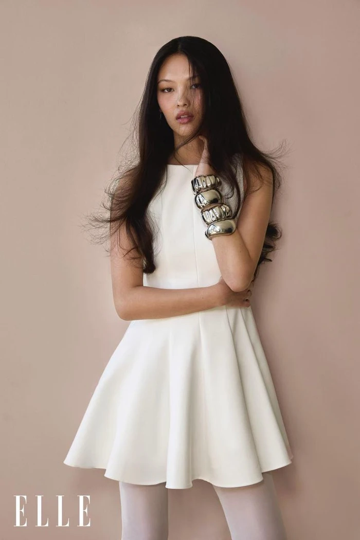 Model May Li Wescott wearing a white sleeveless flared mini dress and chunky silver cuff bracelets against a warm studio backdrop.